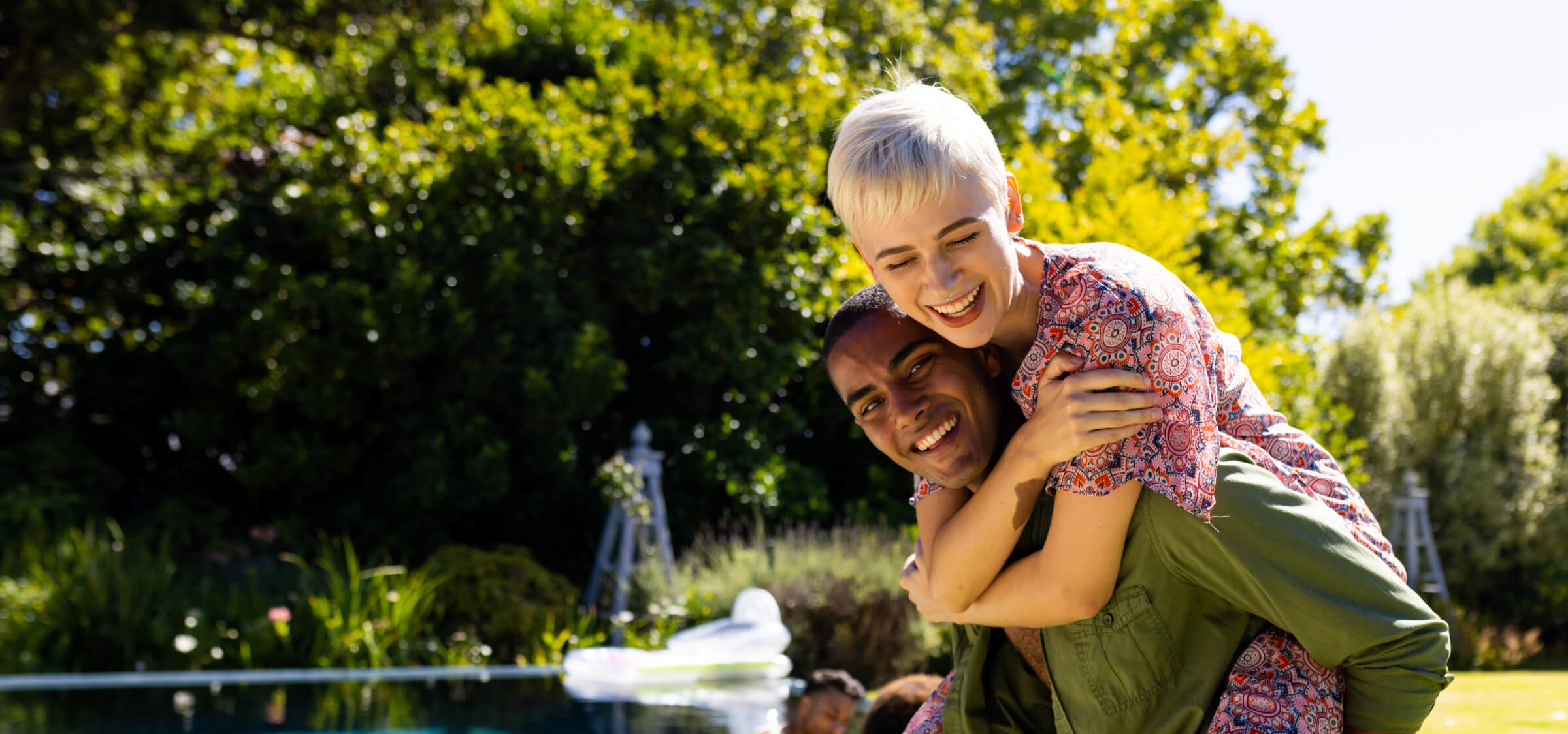 A woman with short blonde hair smiles while giving a piggyback ride to a smiling man near a pool outdoors on a sunny day.