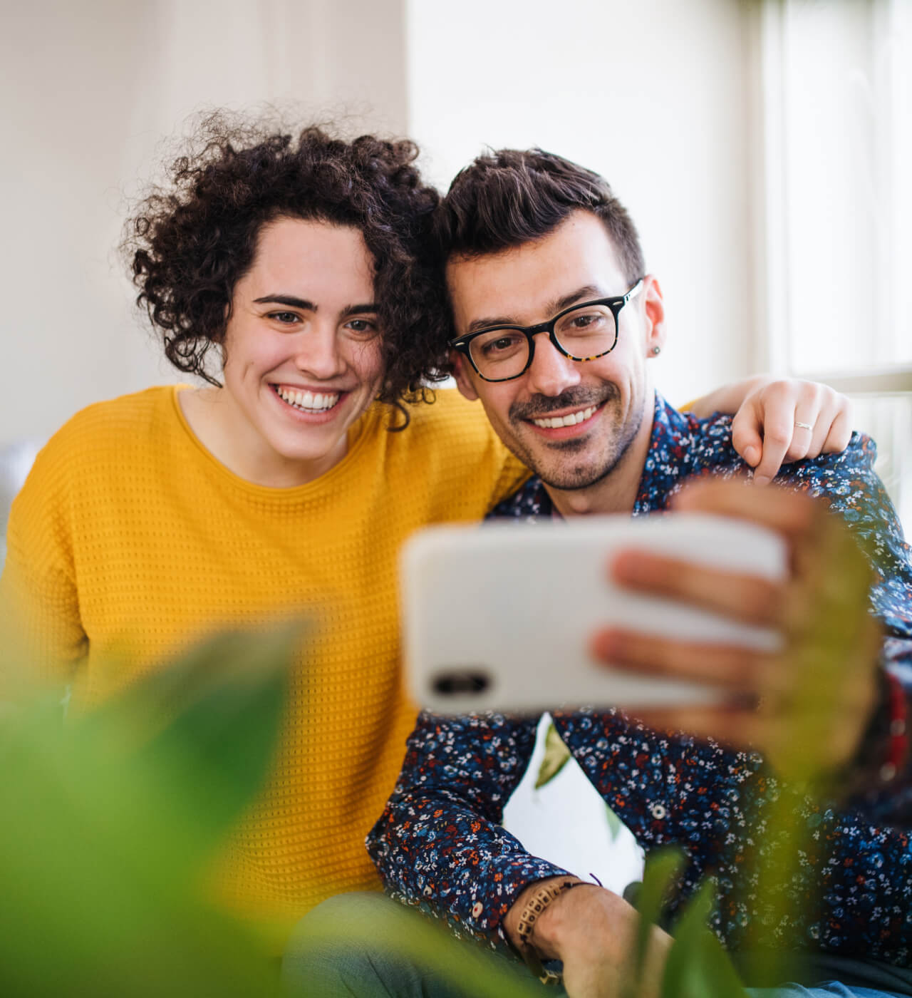 Two people sitting close together, smiling, and taking a selfie with a smartphone. One wears a yellow sweater, the other a patterned blue shirt and glasses. Green foliage obscures part of the foreground.