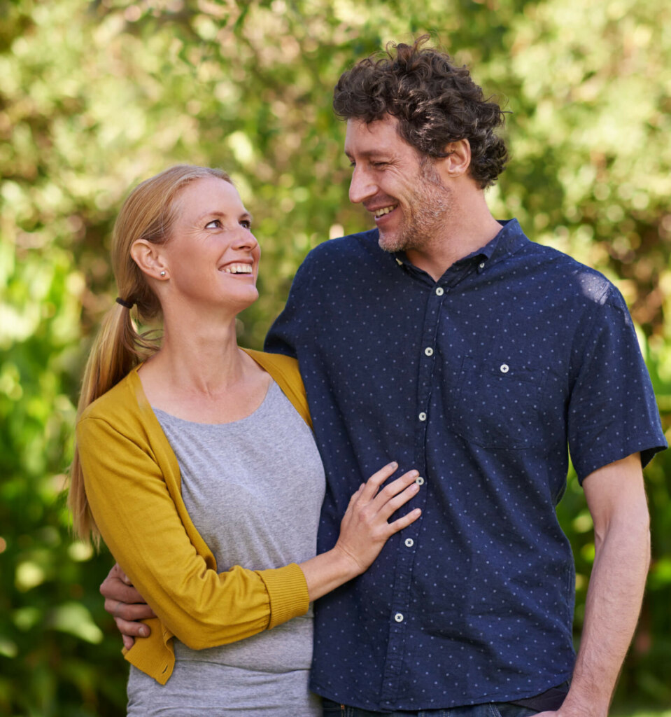 A man and woman stand closely together outside, smiling at each other. The woman wears a yellow cardigan and grey shirt, while the man wears a navy blue button-up shirt with small white dots.