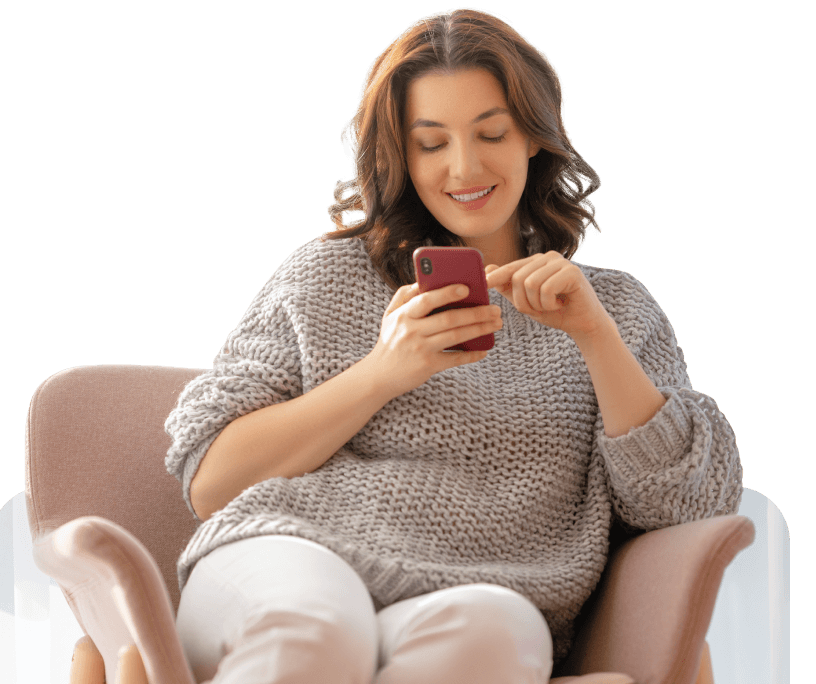 A woman sitting on a chair, wearing a grey sweater and white pants, looking at her smartphone with a smile.