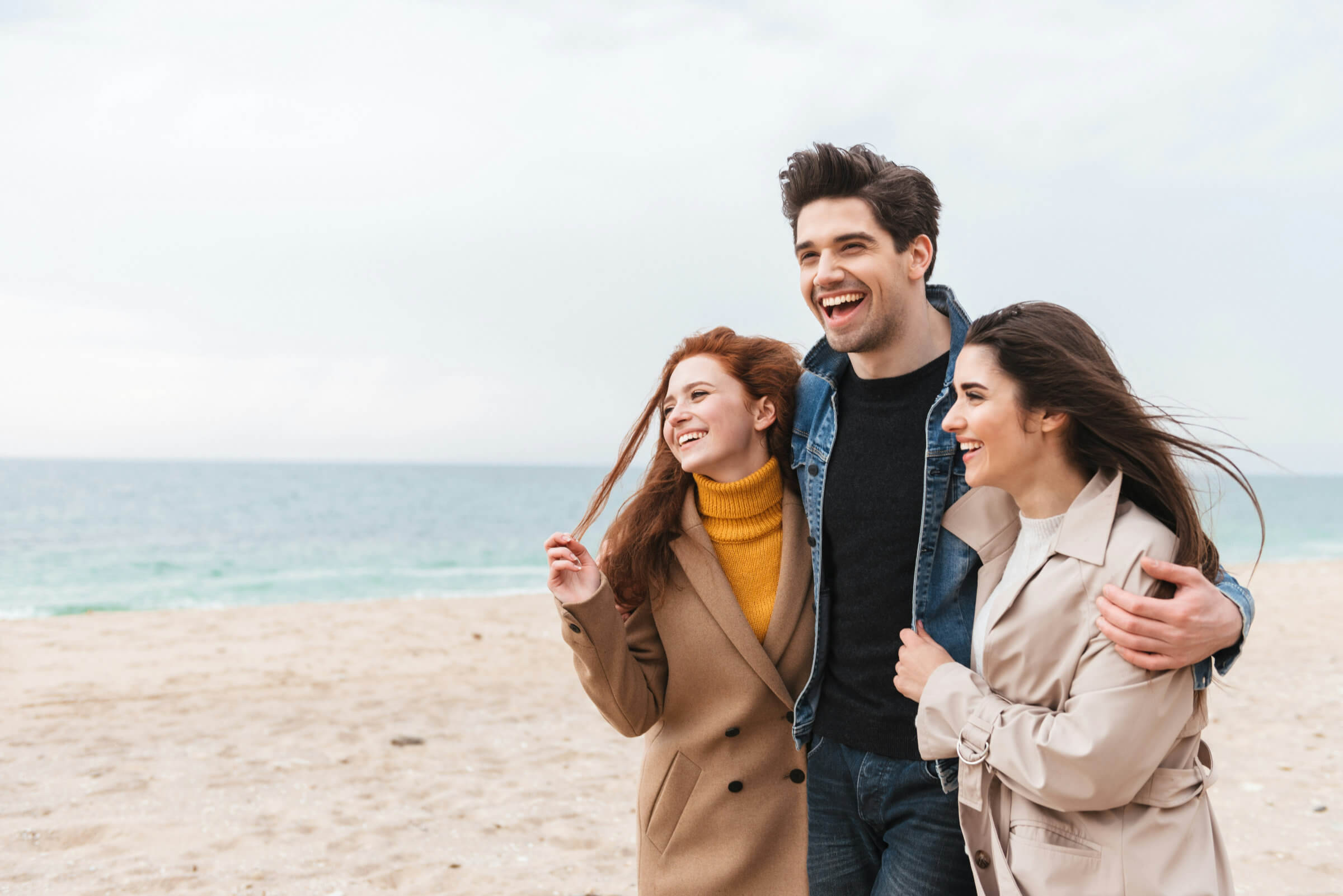 Three people walking together on a beach, smiling and laughing, bundled up in coats on a cloudy day.