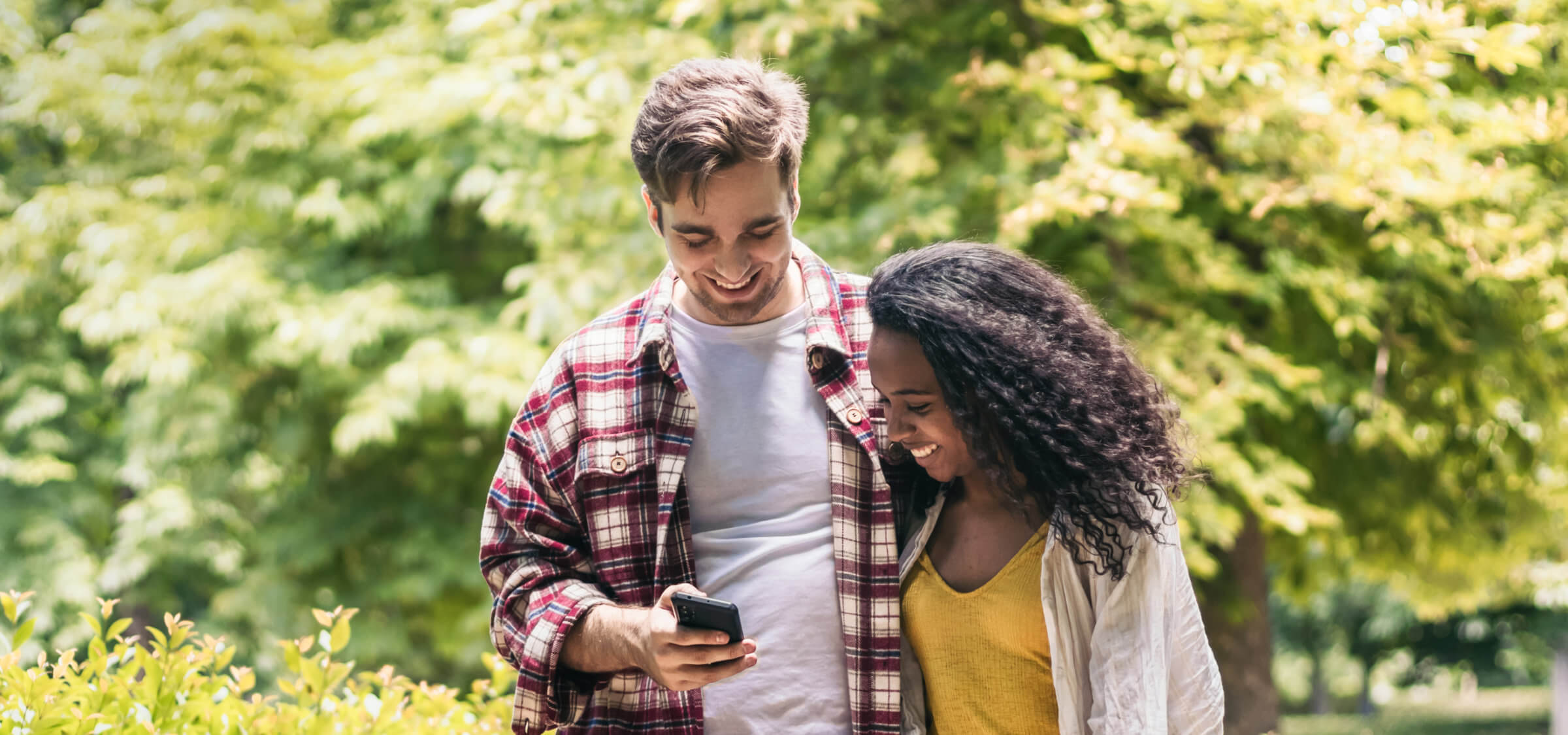 Two people are walking outdoors while looking at a smartphone. The person on the left is wearing a plaid shirt, and the person on the right is wearing a yellow top. Greenery is visible in the background.