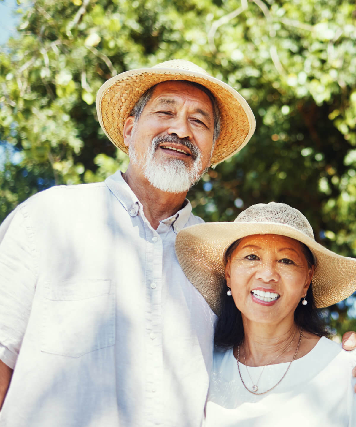 An elderly couple stands outdoors, smiling at the camera. Both are wearing straw hats and light-colored clothing. Trees with green leaves are visible in the background.