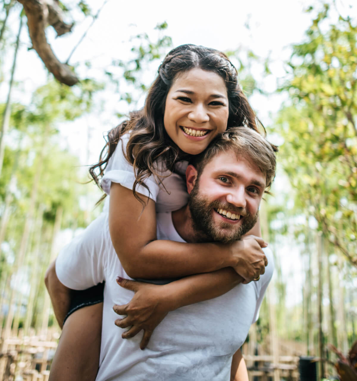 A woman smiles while piggybacking on a smiling, bearded man's back. They are outdoors with trees in the background.