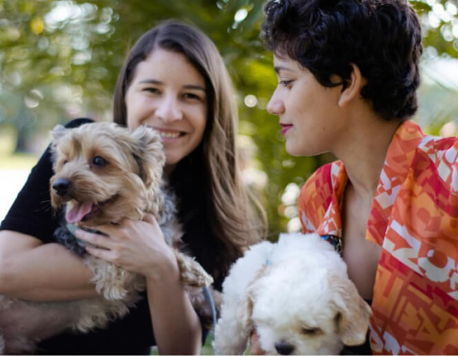 Two individuals are outdoors, each holding a dog. One person has long hair and is holding a small brown dog while smiling. The other person has short hair and is holding a small white dog.