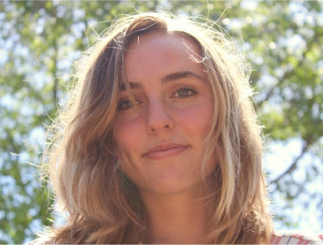 A young person with long, light brown hair smiles slightly while standing outdoors with foliage and sunlight in the background.