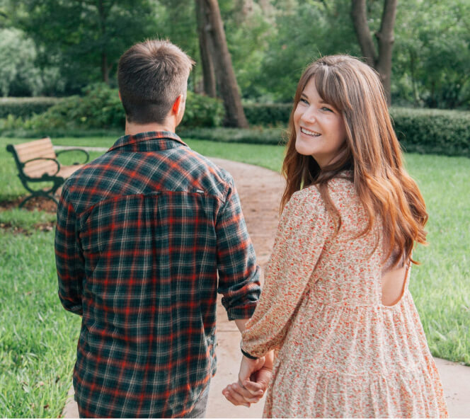 A couple walks hand-in-hand on a park path, with the woman smiling and looking back while the man faces forward. A bench and greenery are visible in the background.