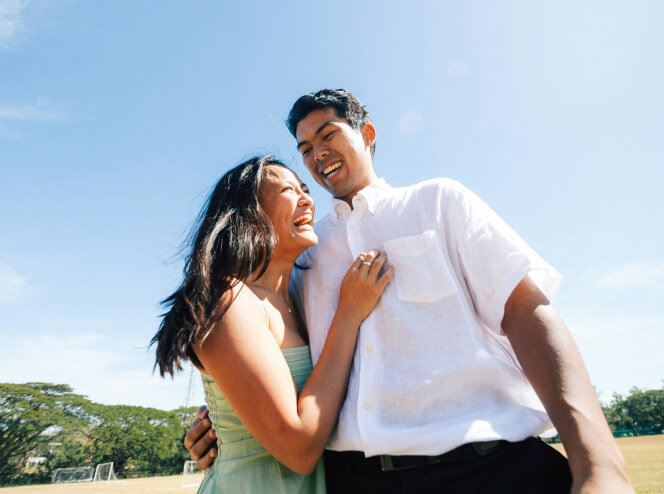 A man and woman stand outside, smiling and laughing. The sky is clear and blue, with some trees visible in the background.