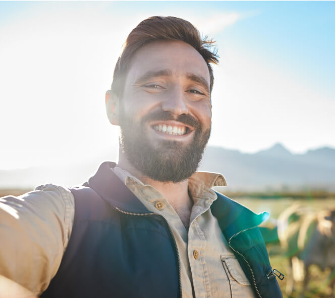 A man with a beard and mustache smiles at the camera while standing outdoors. He is wearing a beige shirt and a dark vest, with a sunny landscape and mountains in the background.